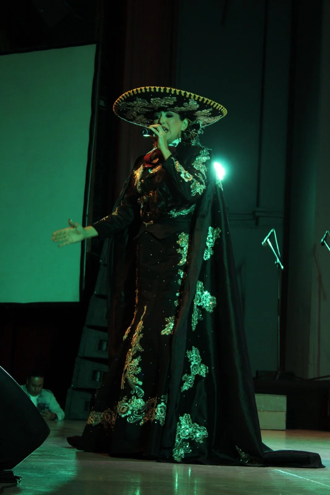 Una mujer vestida con un vestido tradicional negro con bordados dorados, lleva un sombrero grande y sombrero de charro, y canta en un micrófono en un escenario con iluminación verde.