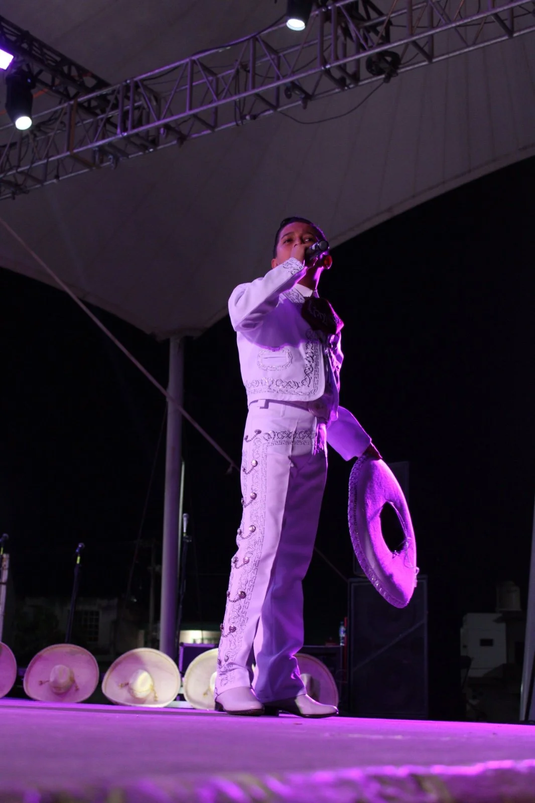 Un niño vestido con traje de mariachi blanco en un escenario, sosteniendo un micrófono y un sombrero grande, en un concierto nocturno con fondo oscuro y luces moradas.