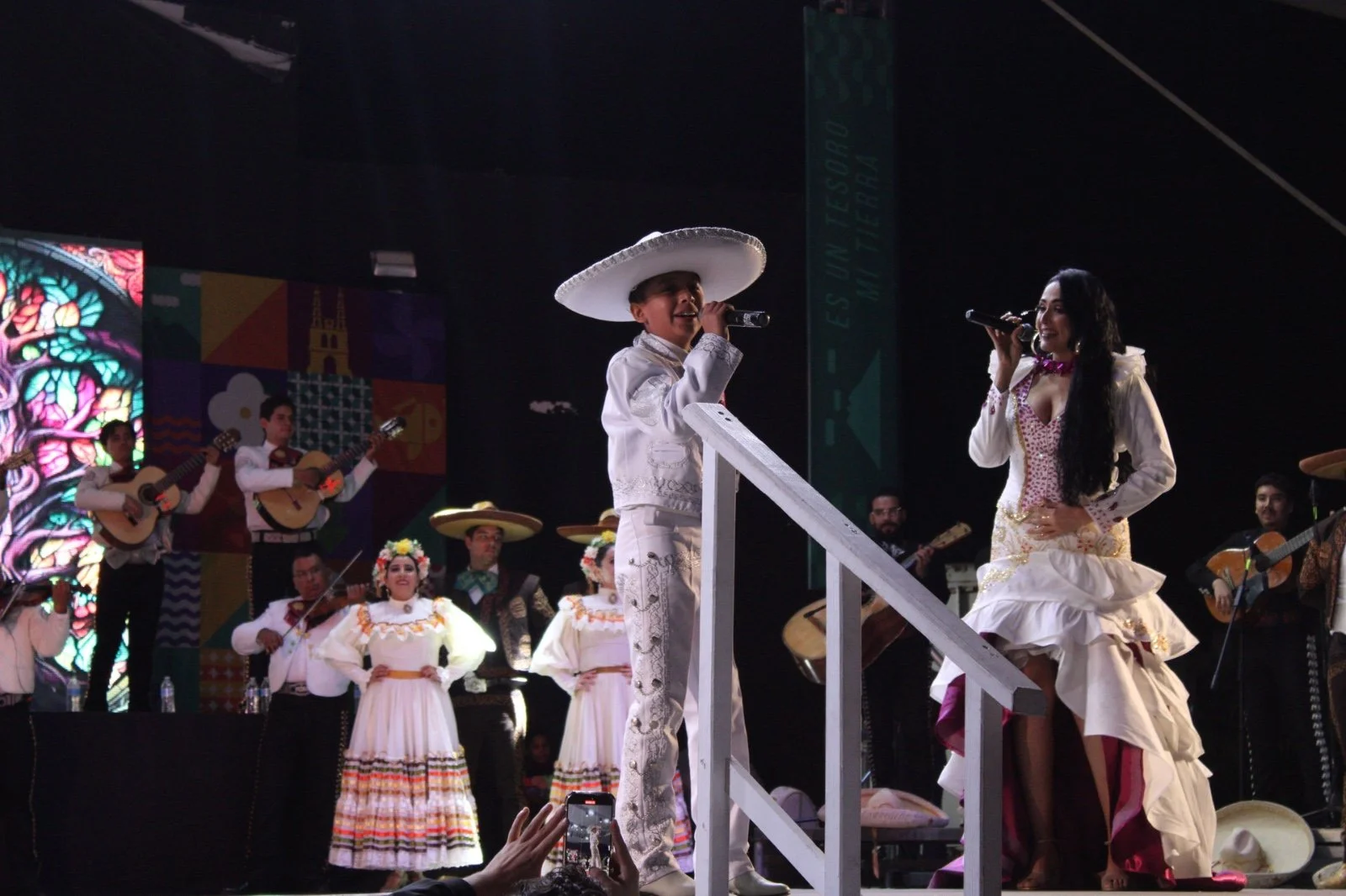 Baile folklórico mexicano con cantantes vestidas con trajes tradicionales y músicos tocando guitarras en el escenario.