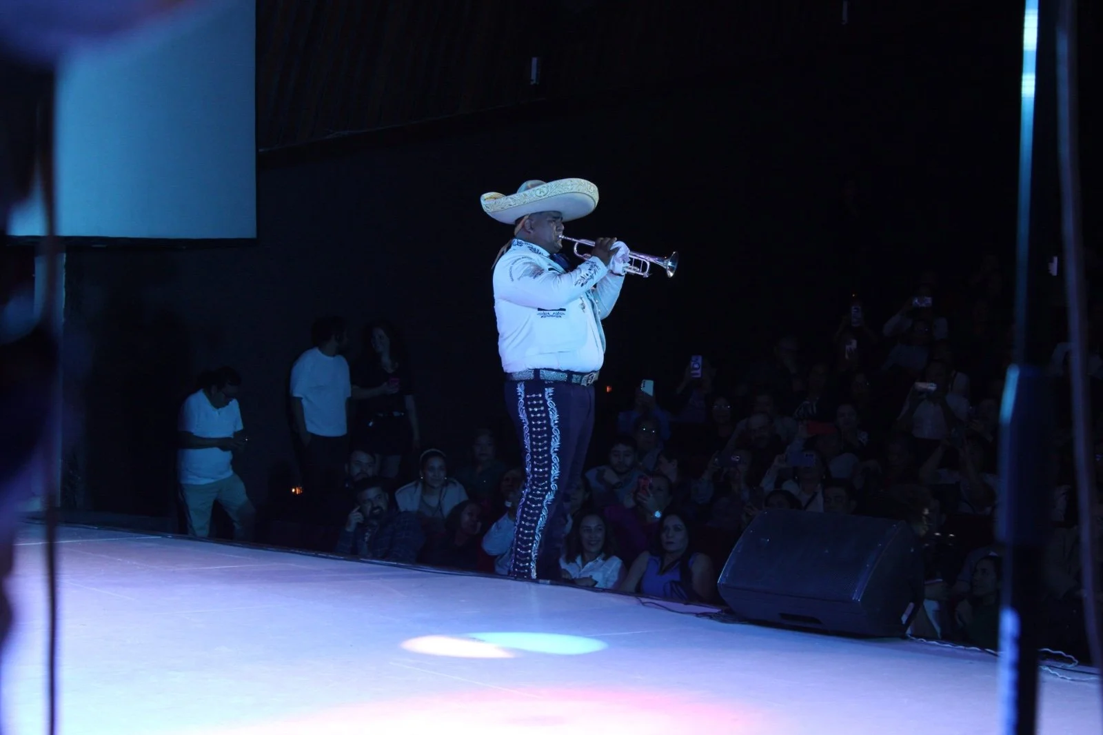 Hombre vestido con traje tradicional mexicano, sombrero grande y tocando la trompeta en un escenario. Audiencia observando en la oscuridad.