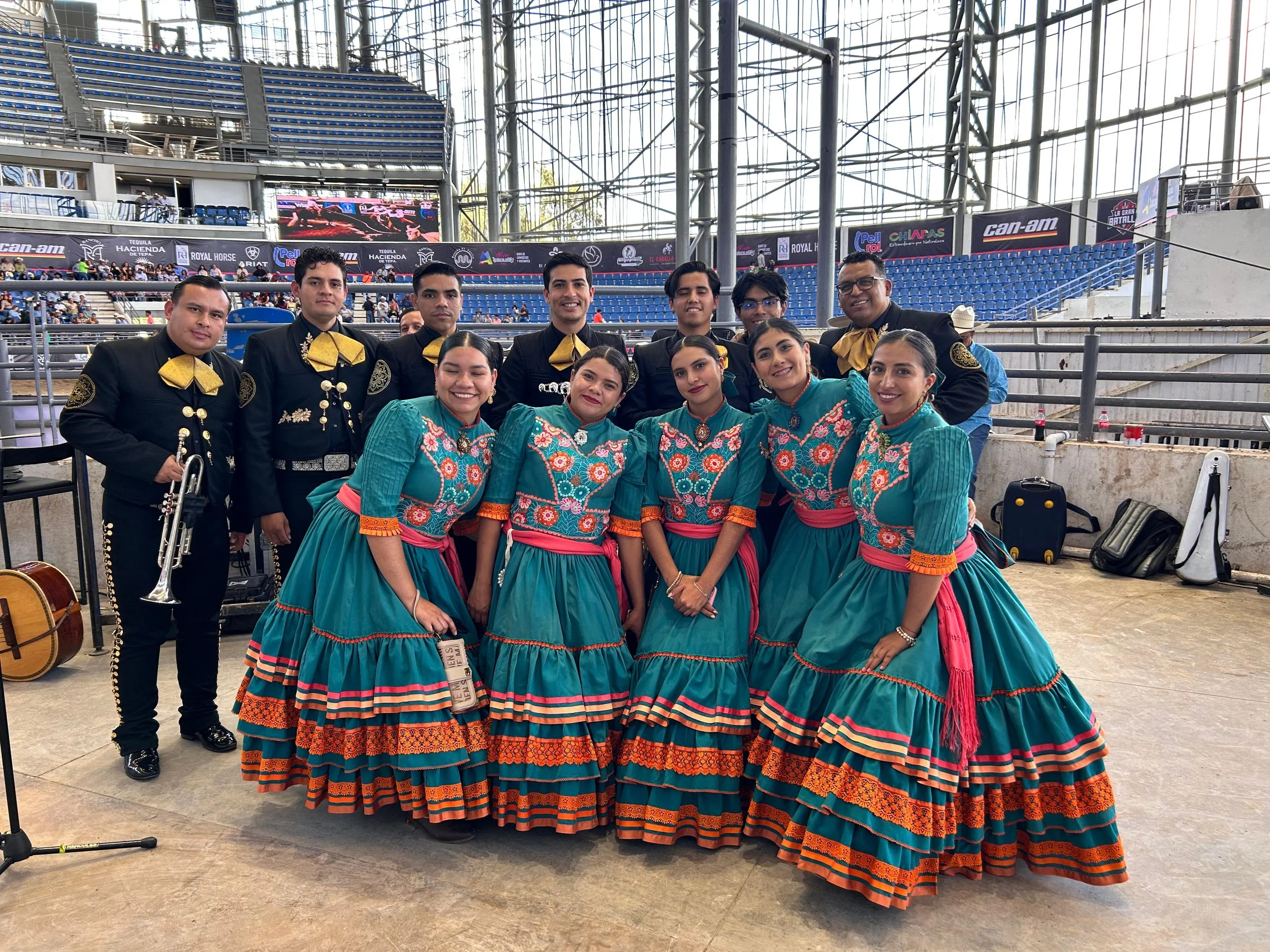 Grupo de bailarines con trajes tradicionales mexicanos posando en un estadio, hombres con trajes negros y mujeres con vestidos azules con detalles en naranja y rosa.