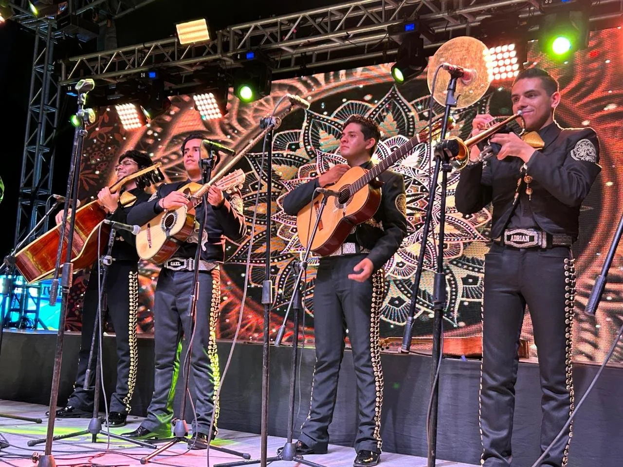 Cuatro músicos de mariachi en el escenario tocando guitarras y trompetas, vestidos con trajes tradicionales negros con detalles dorados, con fondo de pantalla colorido y decoración.