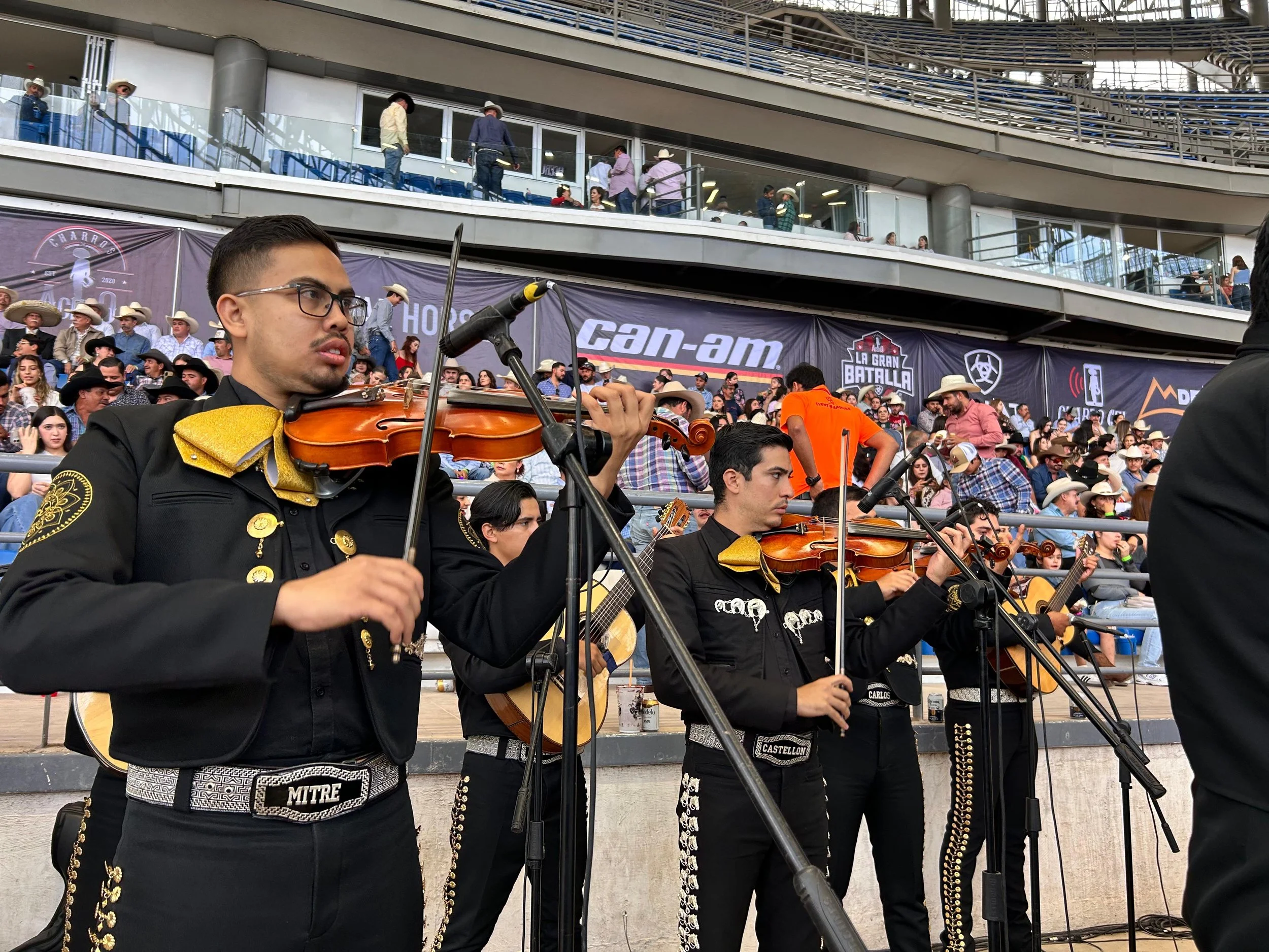 Grupo de músicos de mariachi tocando instrumentos en un escenario con audiencia en el fondo.