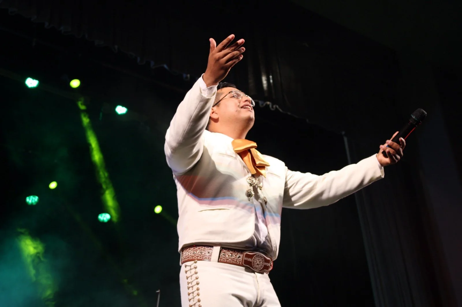 Hombre con traje blanco y accesorios tradicionales mexicanos, sosteniendo un micrófono, en escenario con luces verdes y negras.