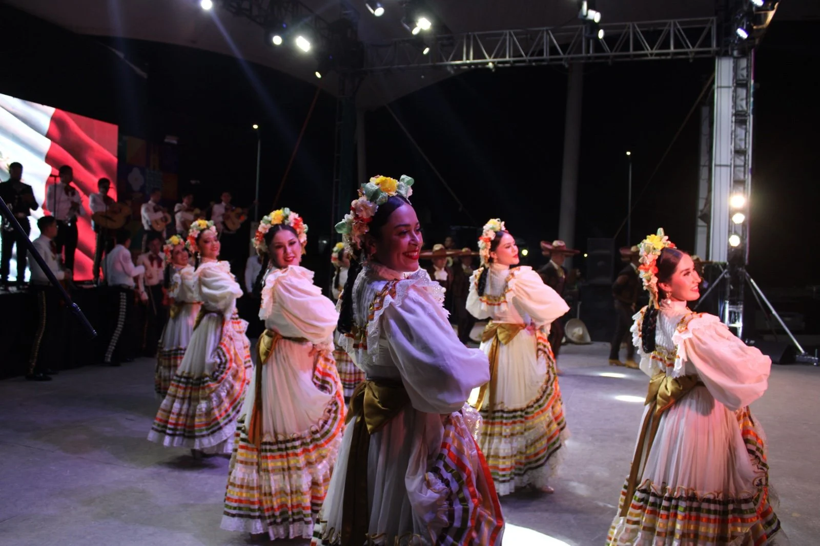 Grupo de mujeres en vestidos tradicionales de colores con flores en la cabeza, bailando en un escenario con músicos en el fondo, durante una celebración cultural nocturna.
