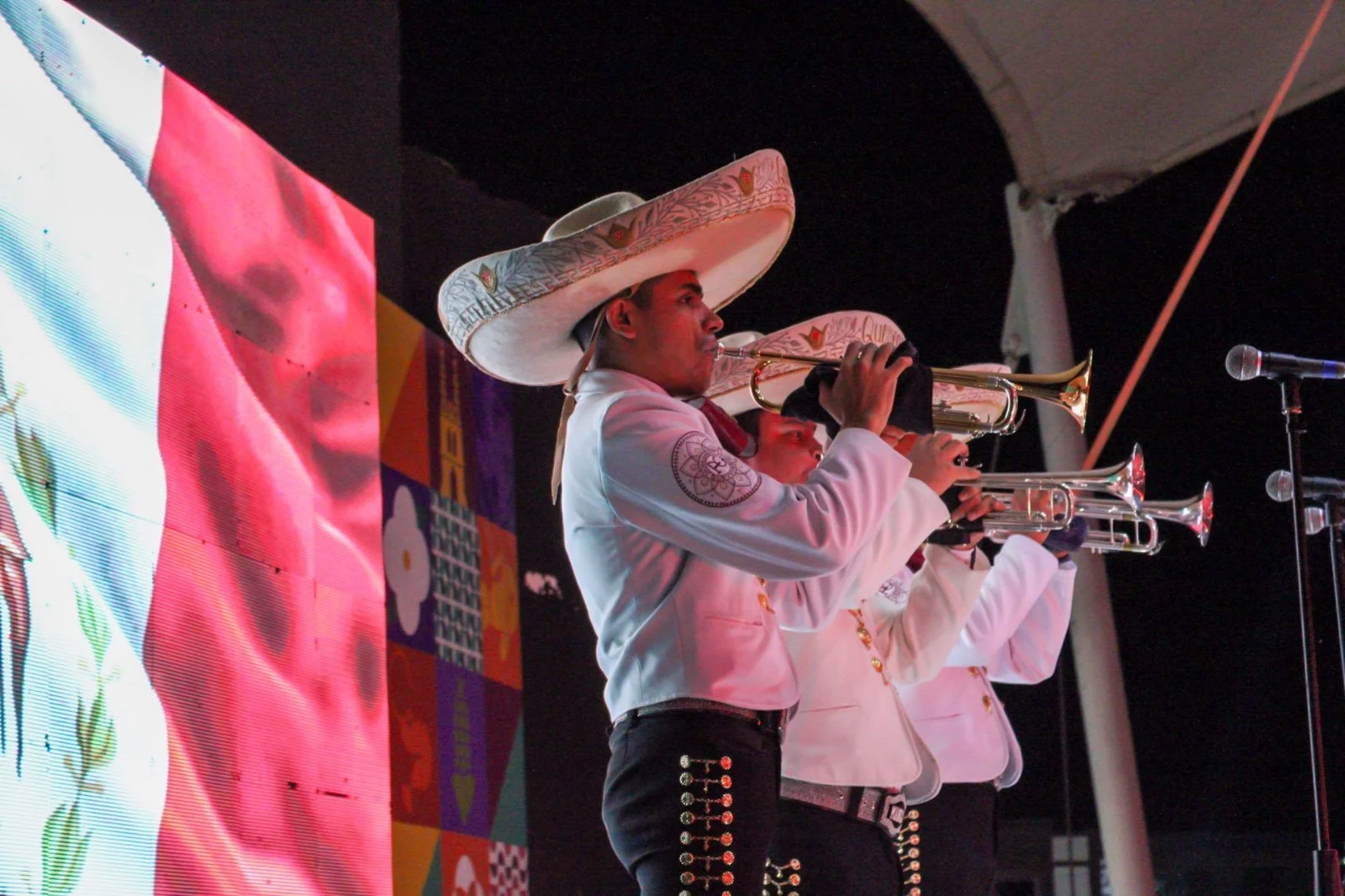 Tres músicos tocando trompetas en un escenario, usando sombreros grandes y trajes blancos con detalles negros y dorados.