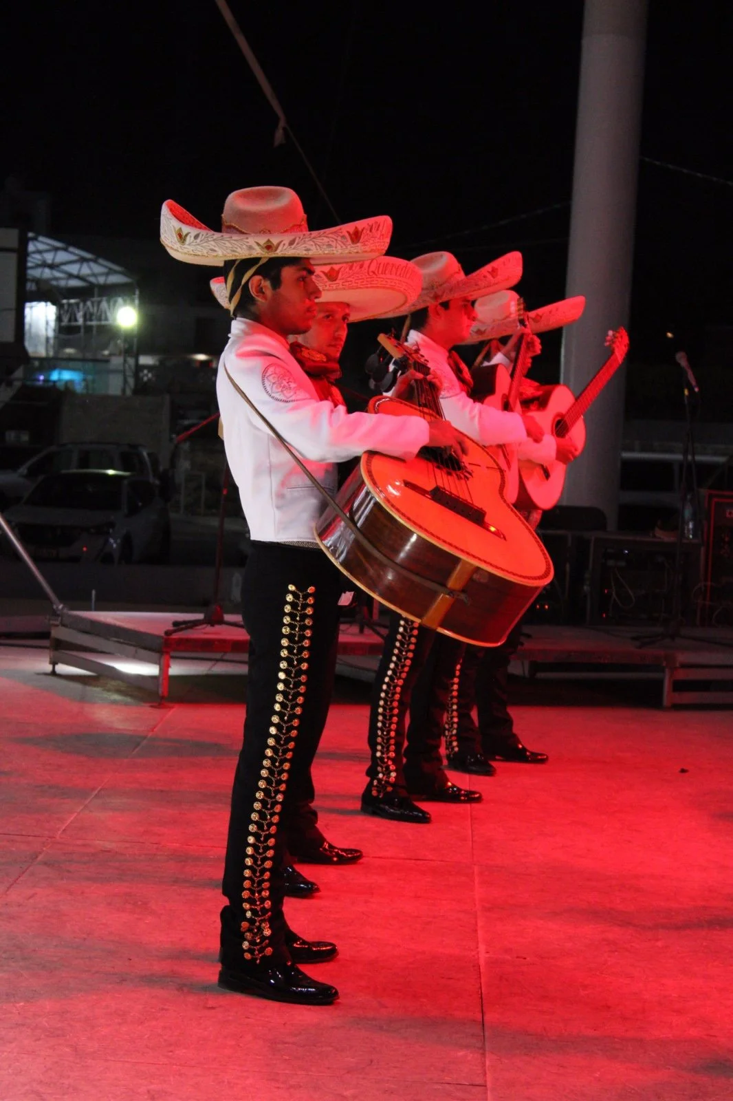 Grupo de músicos mexicanos vestidos con trajes tradicionales, tocando guitarras y violines en una actuación nocturna con sombreros de mariachi.