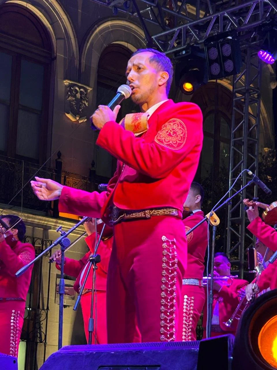 Hombre vestido con traje tradicional mexicano rojo, tocando un violín en un escenario iluminado con luces moradas y doradas, con otros músicos de fondo también vestidos de rojo.