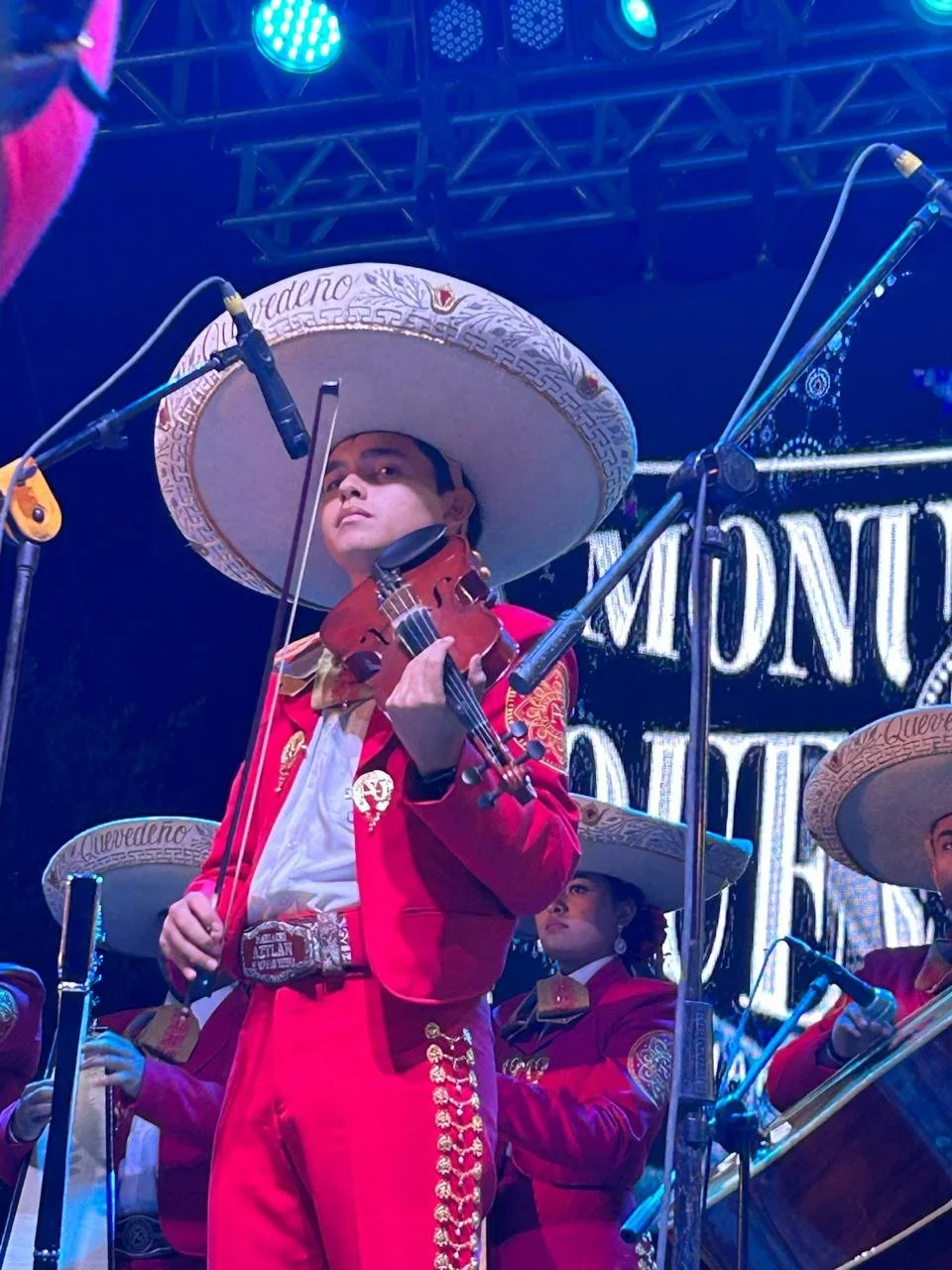 Un grupo de músicos vestidos con trajes tradicionales mexicanos rojos y sombreros grandes y decorados, tocando instrumentos en un escenario con luces azules y fondo con letras grandes.