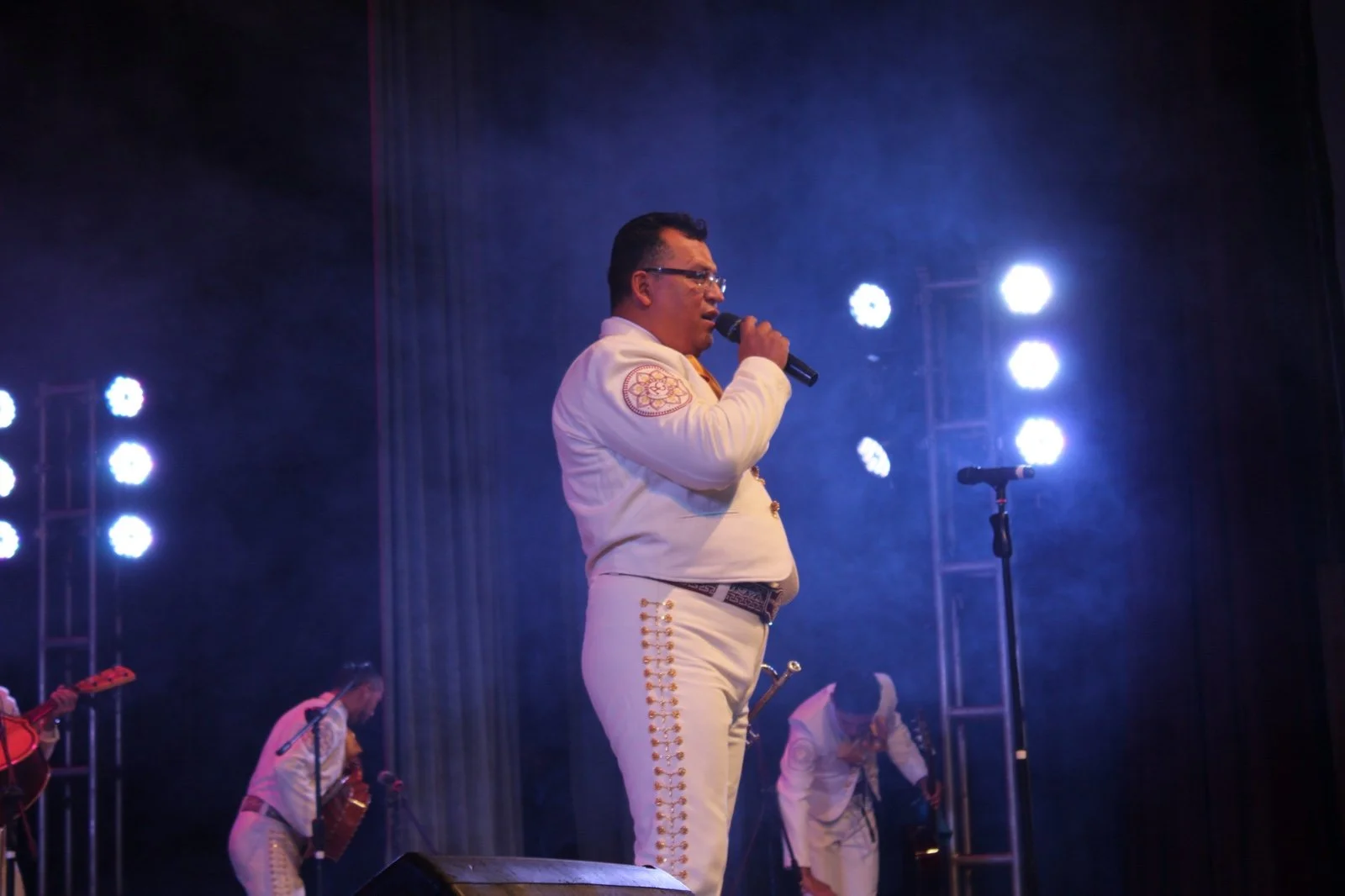 Hombre vestido con traje tradicional mexicano cantando en un escenario con micrófono, con otros músicos en el fondo y luces brillantes.