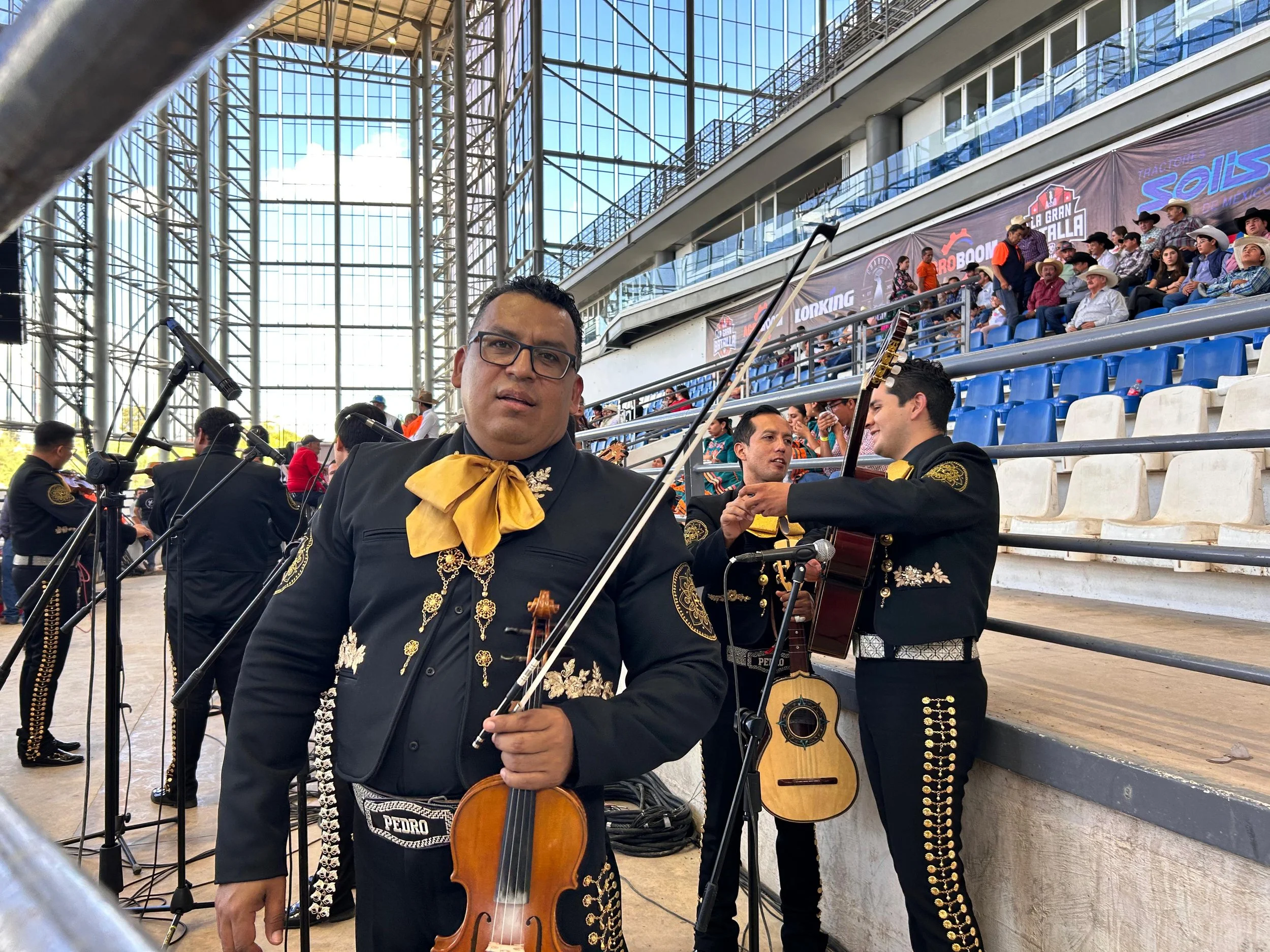 Fotografía de un grupo de músicos mexicanos disfrazados de charros, con instrumentos musicales, en un evento cultural en una plaza cubierta con espectadores en las gradas.