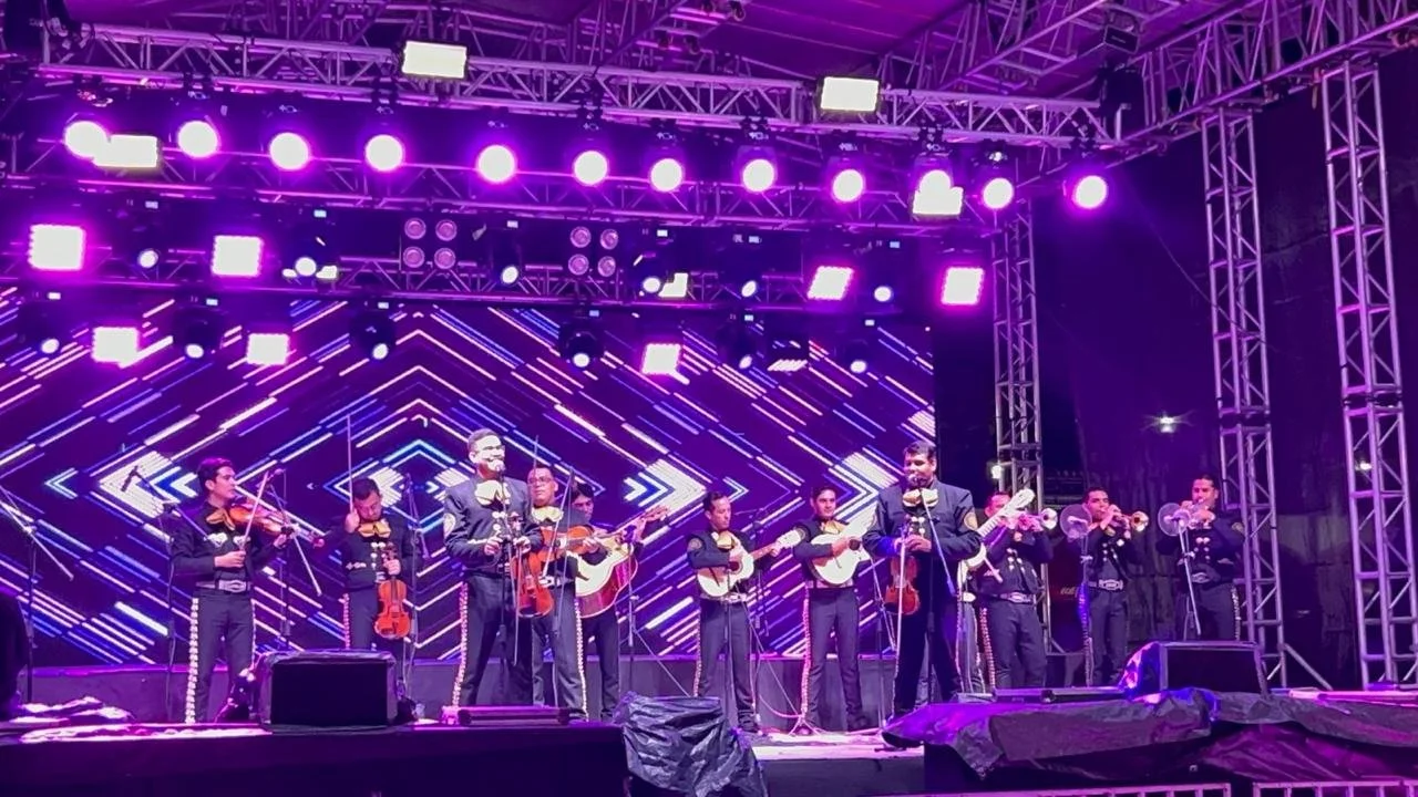 Conjunto musical en escenario con instrumentos de cuerda y trompetas, fondo de luces y pantallas en colores violeta y azul.