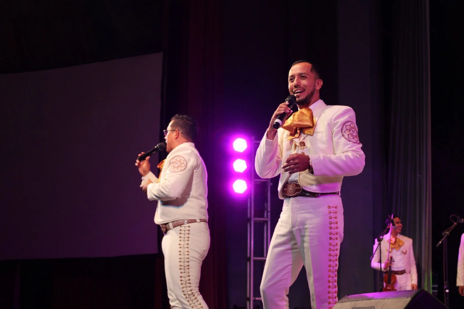 Un grupo de mariachis en el escenario, tocando y cantando, vestidos con trajes tradicionales blancos con detalles dorados y amarillos.