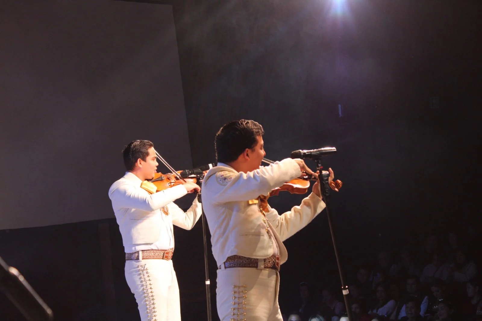 Tres músicos tocando violín en un escenario con audiencia observando.