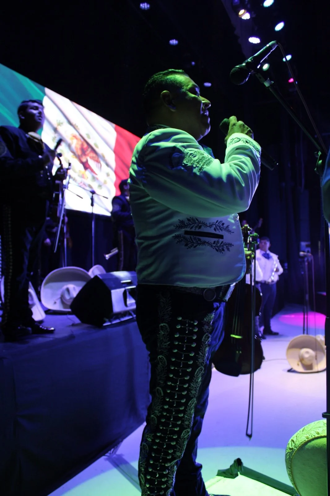 Un grupo de músicos en un escenario, con un hombre en primer plano que canta o habla frente a un micrófono, con un fondo que muestra la bandera de México.