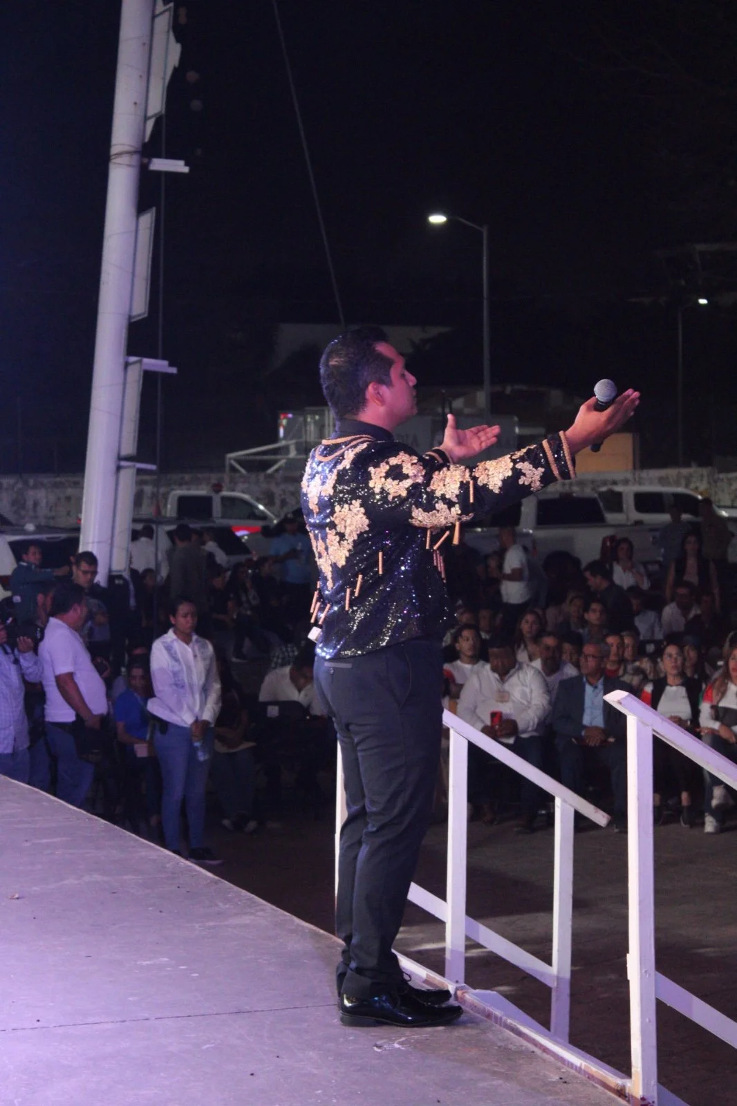 Un hombre en un escenario con un micrófono en mano, vestimenta elegante con detalles dorados, dirigiéndose a una audiencia durante un evento nocturno.