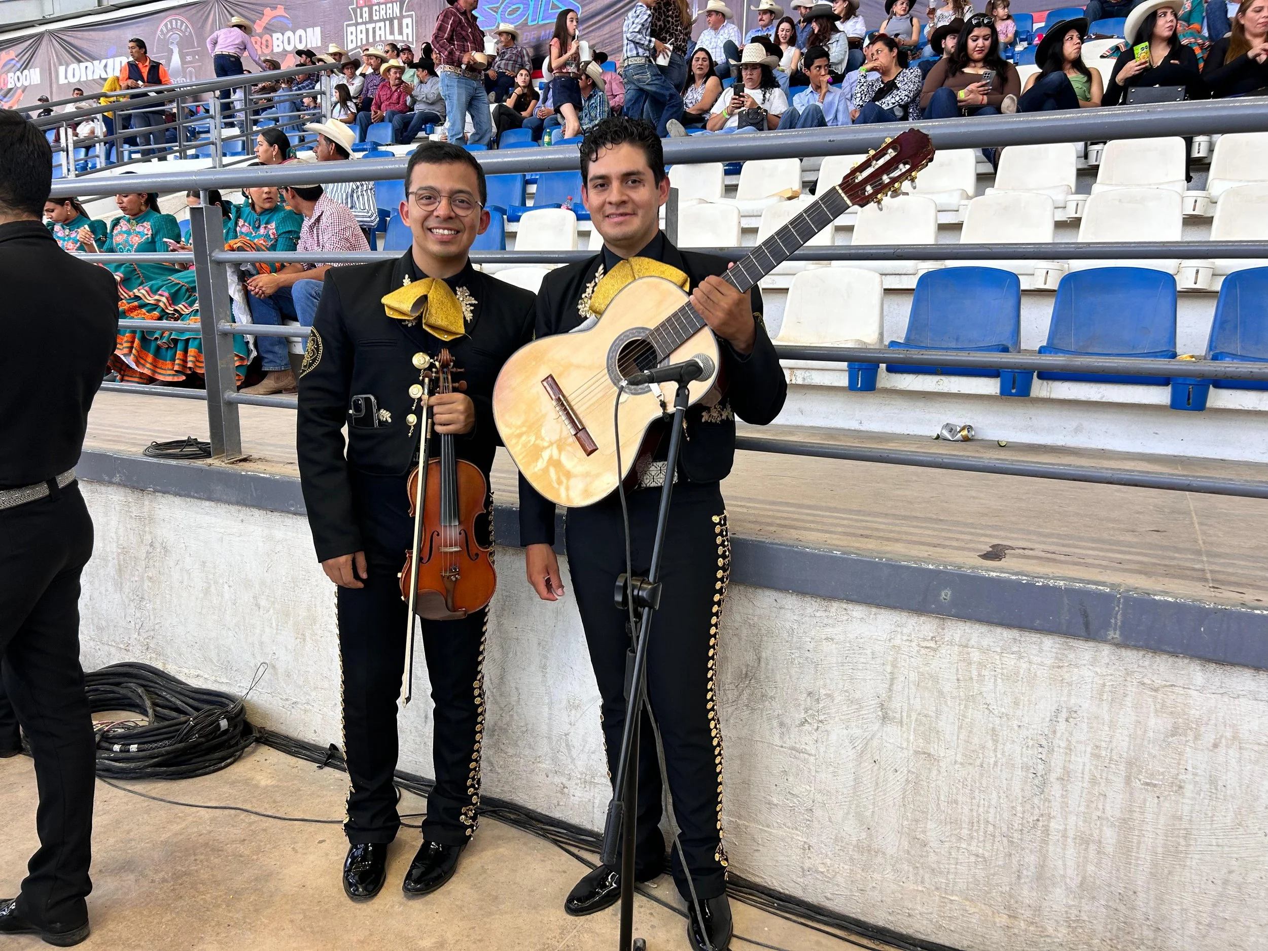 Dos músicos mexicanos con trajes tradicionales, uno con violín y el otro con guitarra, en un evento con público, algunos vestidos con trajes típicos mexicanos.