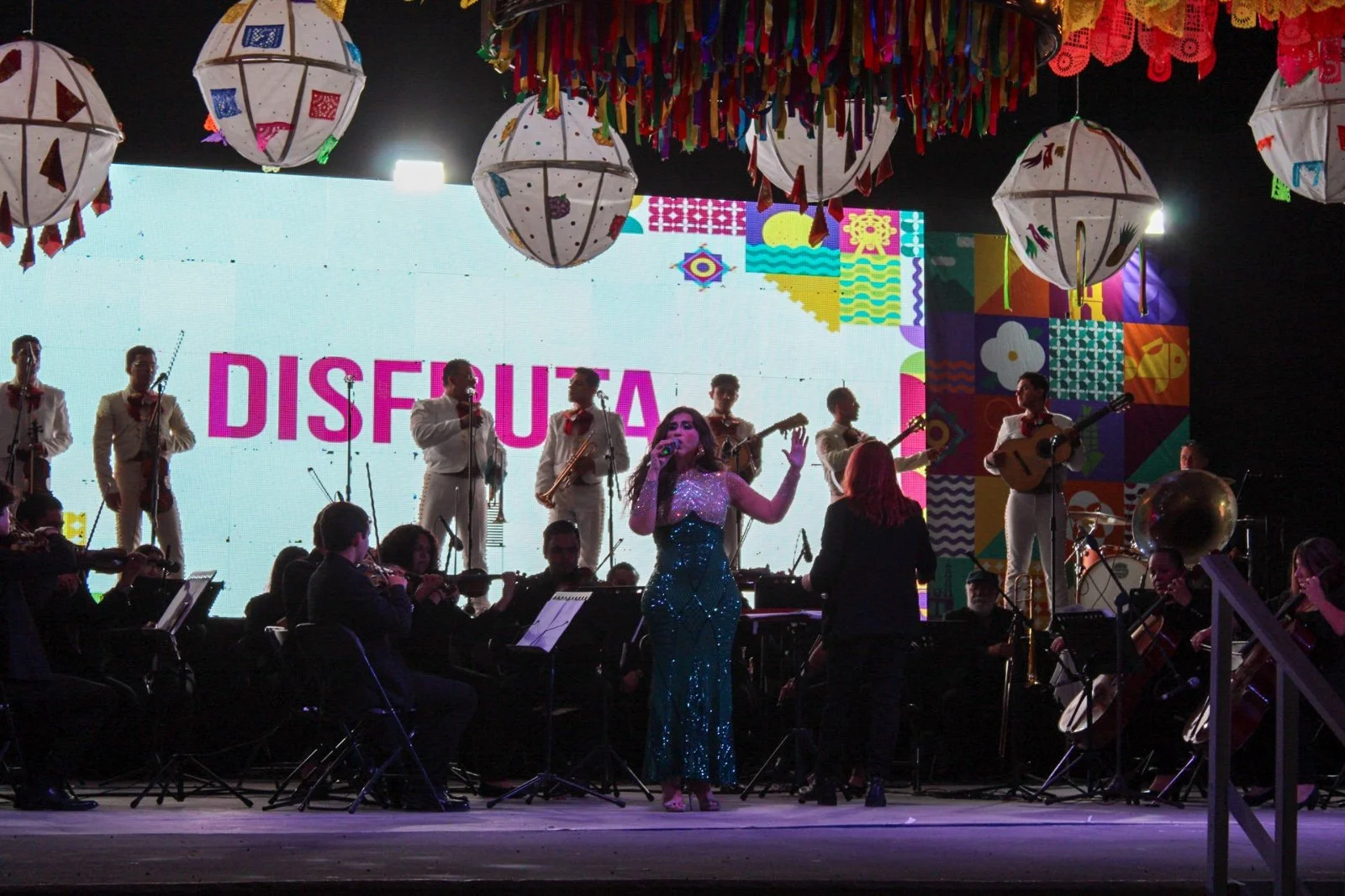 Un escenario con una música en vivo, varias personas tocando instrumentos y cantantes, decorado con faroles de papel y adornos coloridos, con la palabra 'DISFRUTA' en la fondo digital.