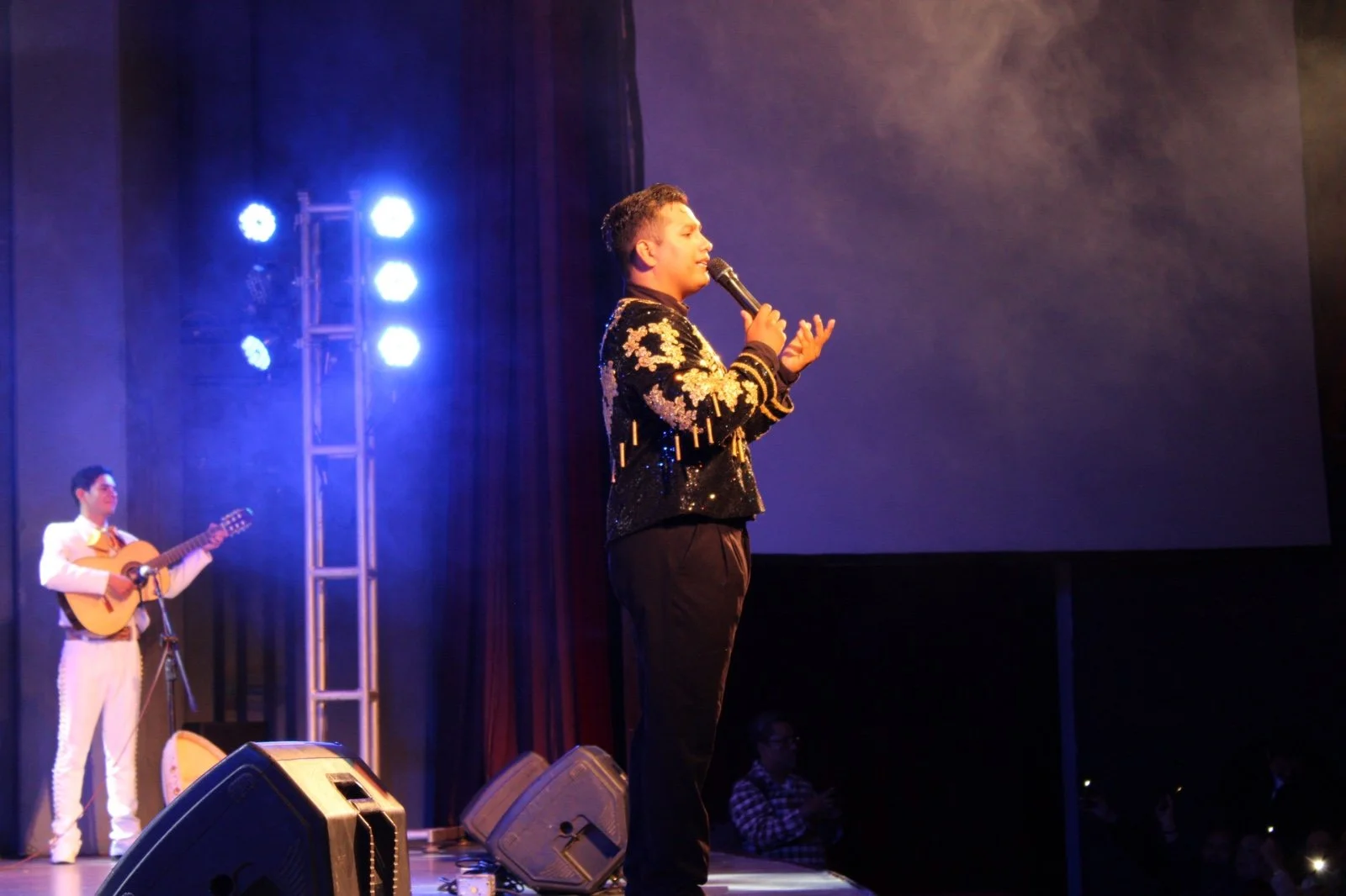 Un cantante en el escenario con un micrófono, vestido con ropa brillante, acompañado por un guitarrista en un espectáculo en un teatro con luces azules.