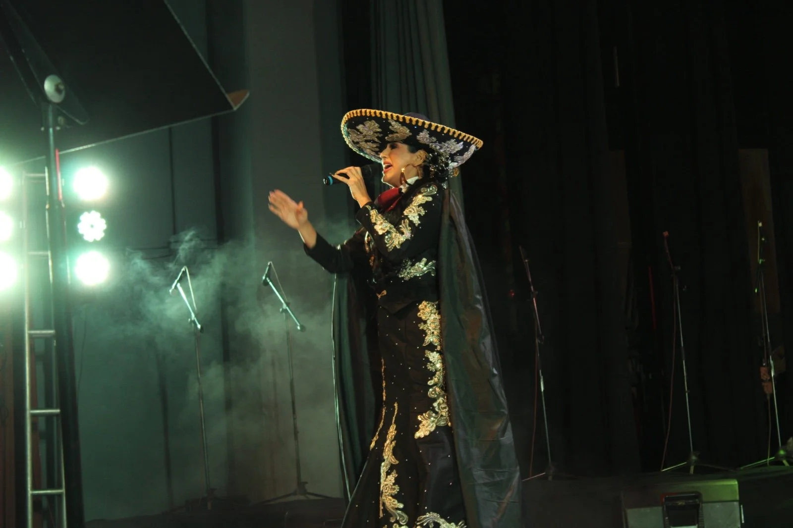 Una mujer vestida con traje tradicional mexicano, con un sombrero grande y adornos dorados, cantando en un escenario con micrófono, luces y humo.
