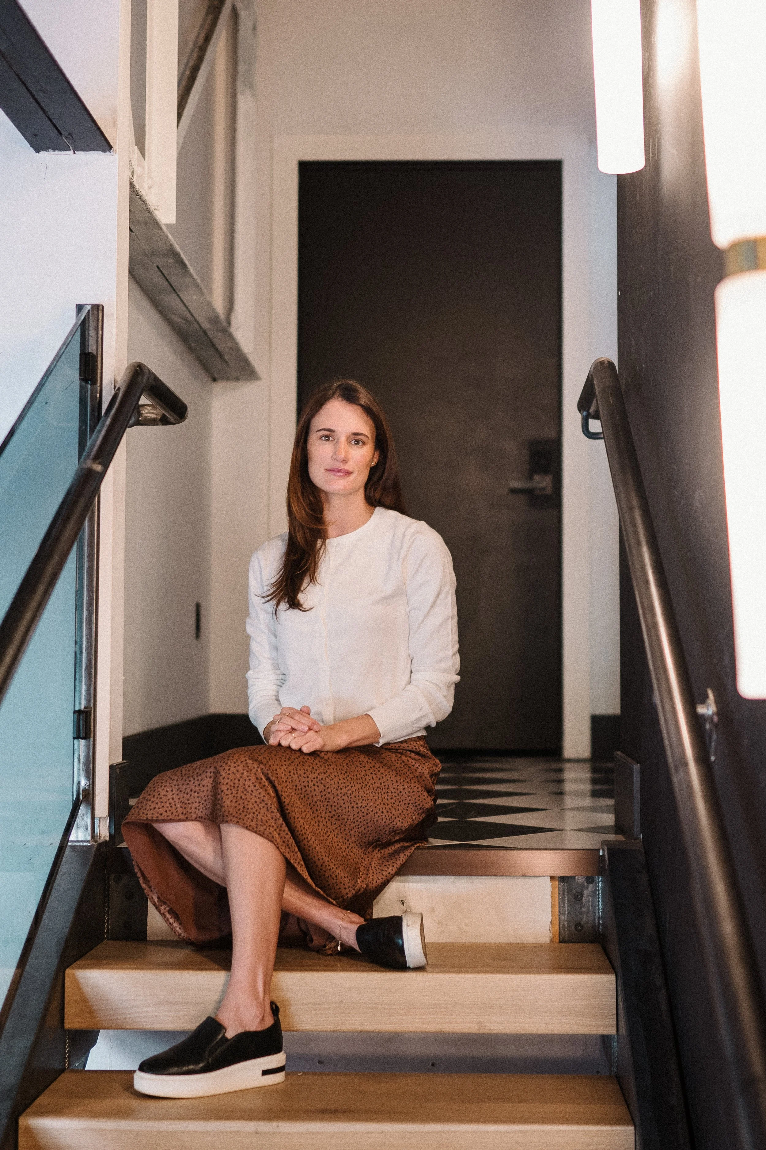A woman sitting on wooden stairs at the entrance of a building with black and white checkered floor, white walls, and a black door in the background.