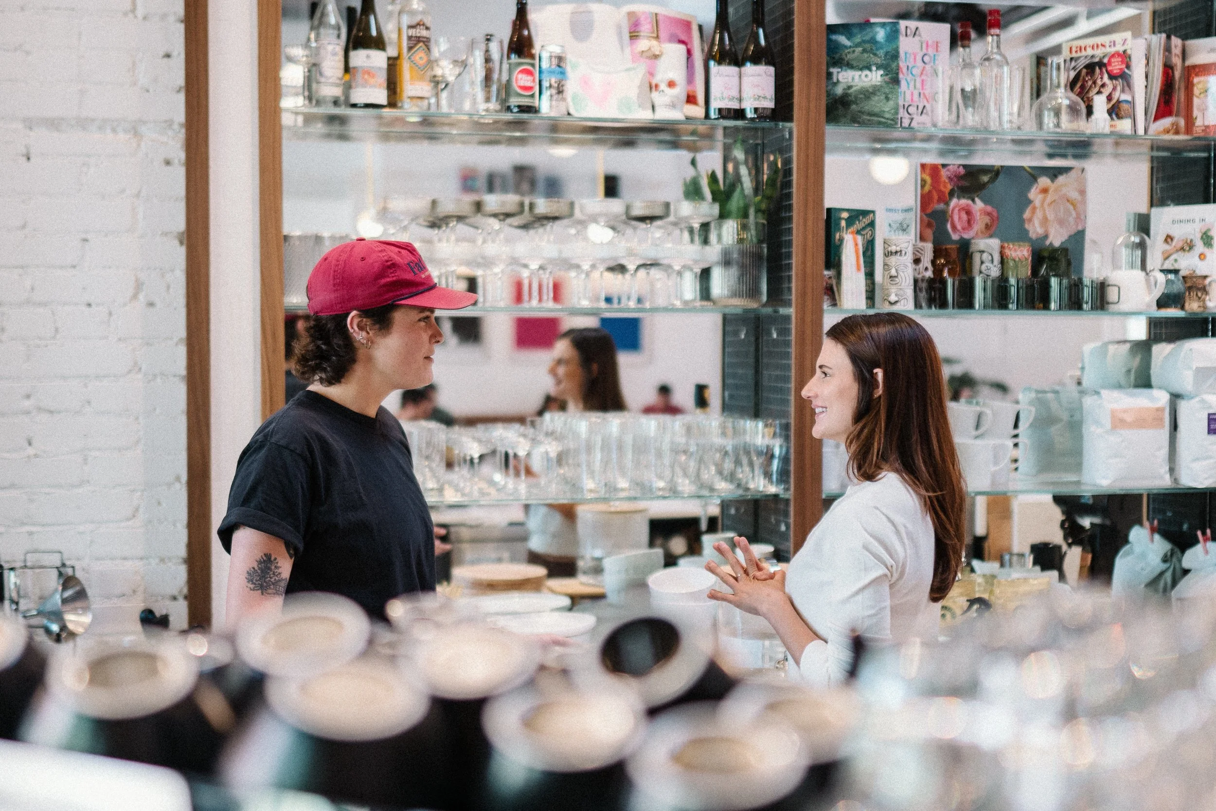 Two women are standing and talking in a cafe or shop. One woman wears a red baseball cap and black shirt, the other wears a white shirt. Behind them are shelves with bottles, glasses, books, and decorative items.