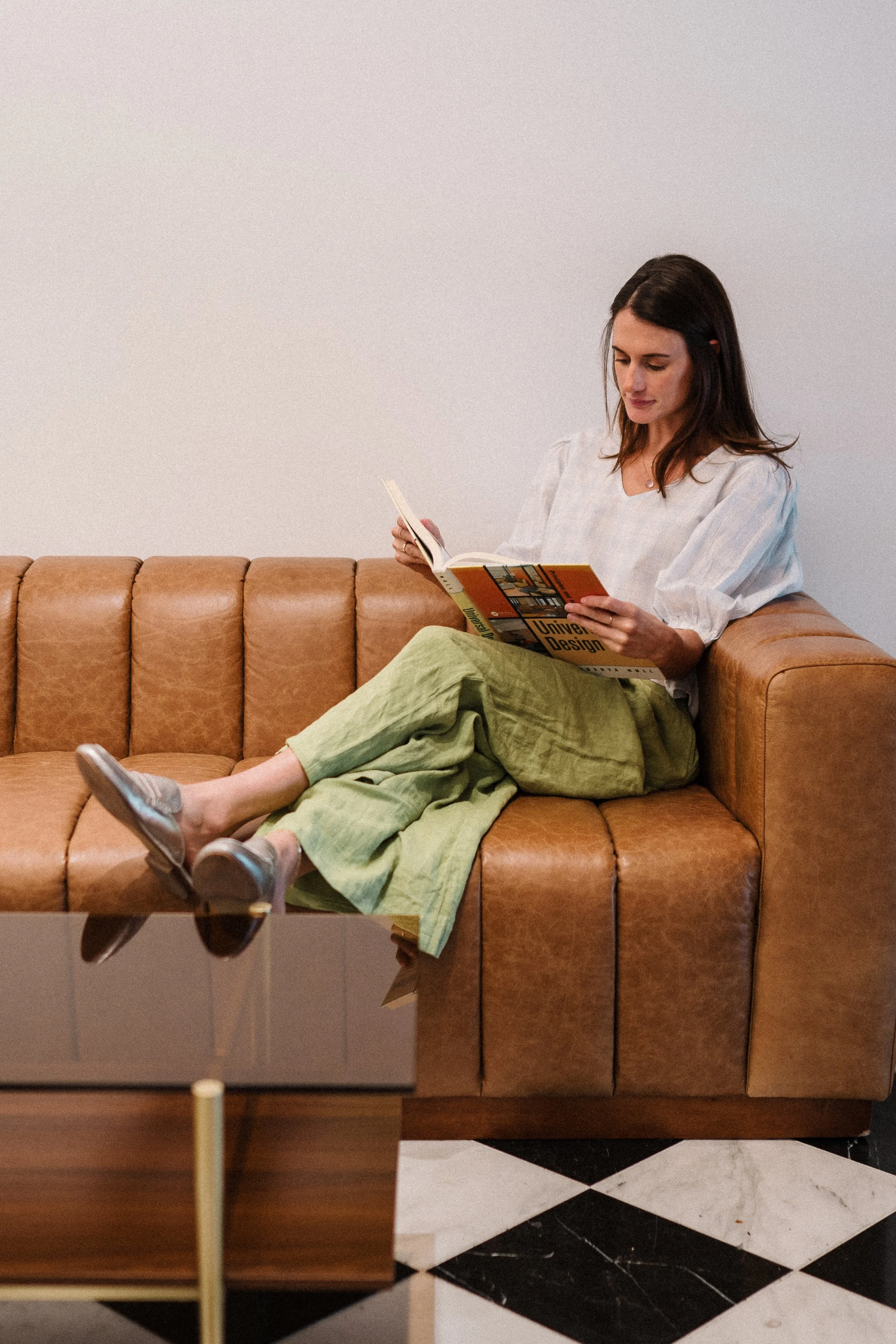 Woman sitting on tan leather sofa reading a book in a modern living room with black and white checkered floor.