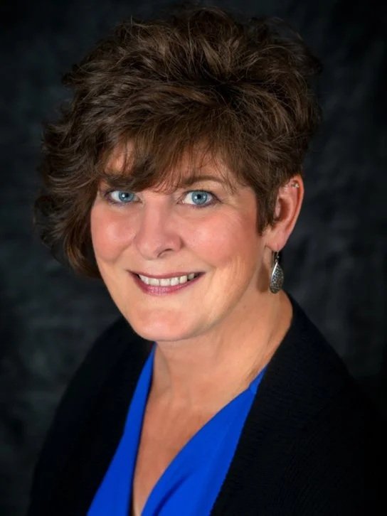 A woman with short, curly brown hair, blue eyes, and light skin, smiling and wearing earrings, a dark blazer, and a blue top against a dark background.