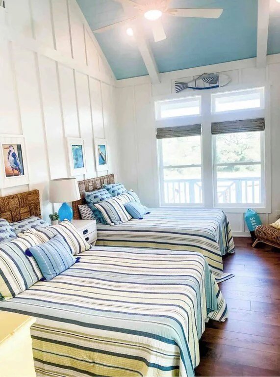 A bright bedroom with two beds, striped bedding in shades of blue, white, and green, and nautical decor. There are three windows with views of water, a ceiling fan with a light, a wicker chair, and seaside-themed artwork on the wall.