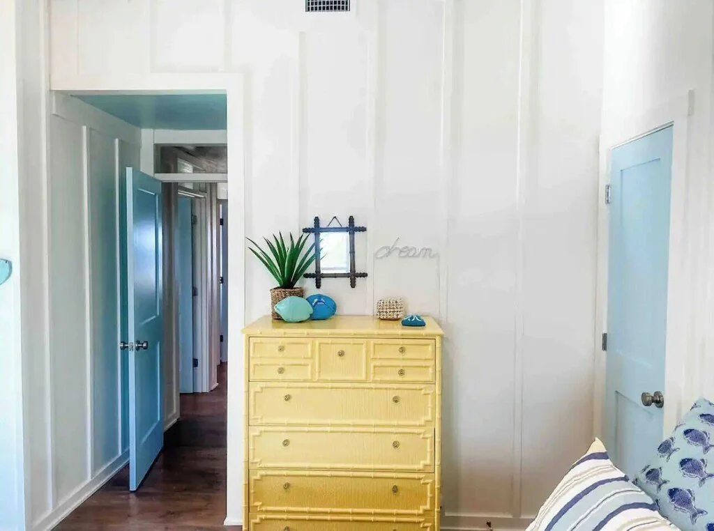 A yellow chest of drawers with decorative items on top, including a potted plant, sea shells, and a small plant. Behind it, a wall decor with the word 'dream' and a small window frame. To the right, part of a bed with patterned pillows. To the left,