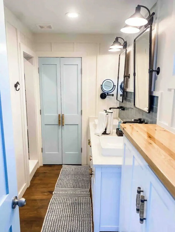 Bathroom with two large mirrors, wall-mounted black lights above mirrors, white farmhouse-style vanity with a vessel sink, wooden countertop on side cabinet, wall-mounted magnifying mirror, black faucet, and a small woven rug on wooden floor.