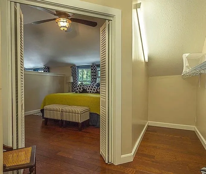 View into a bedroom with a bed, window, and patterned curtains, seen through an open doorway from an adjacent room with a laundry rack.