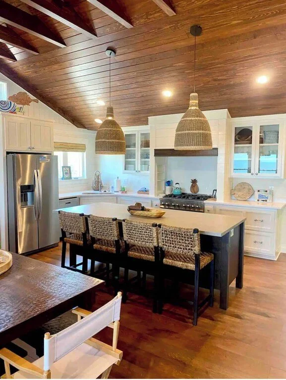 Modern kitchen with white cabinets, stainless steel refrigerator, wooden ceiling, island with four woven chairs, and hanging woven pendant lights.