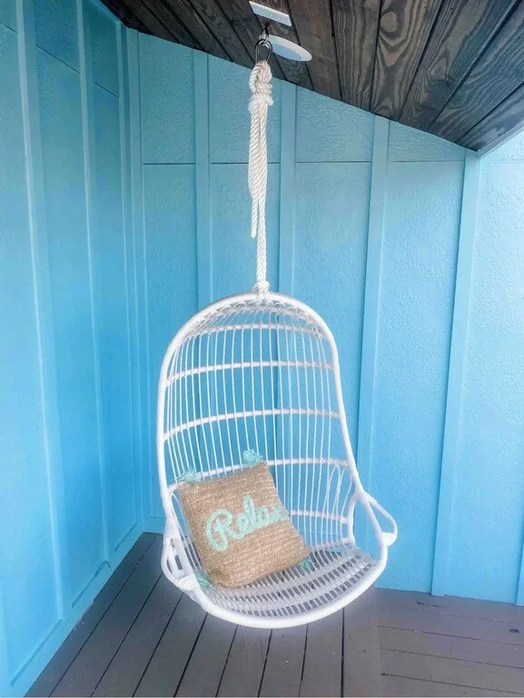 White hanging outdoor swing chair with a beige cushion that says "Relax" on it, set against a blue wooden wall.