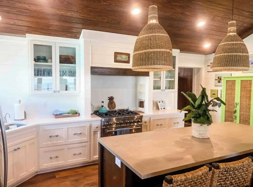 A bright kitchen with white cabinets, a gas stove, wooden ceiling, hanging woven pendant lights, and a kitchen island with plants.