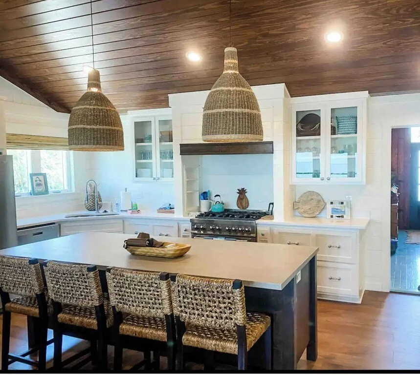 Cozy kitchen with white cabinets, wooden ceiling, and woven pendant lights over a kitchen island with four woven bar stools. Contains a stove, sink, and decorative items.