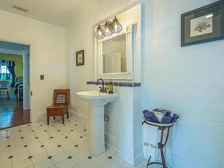 Small bathroom with white walls, a pedestal sink, a mirror above the sink, a light fixture with three bulbs, framed artwork on the walls, and a small table with towels. An open doorway shows a room with green and blue decor.
