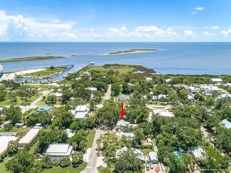 Aerial view of a coastal residential neighborhood with houses and trees near the water, including a marina and islands in the distance.