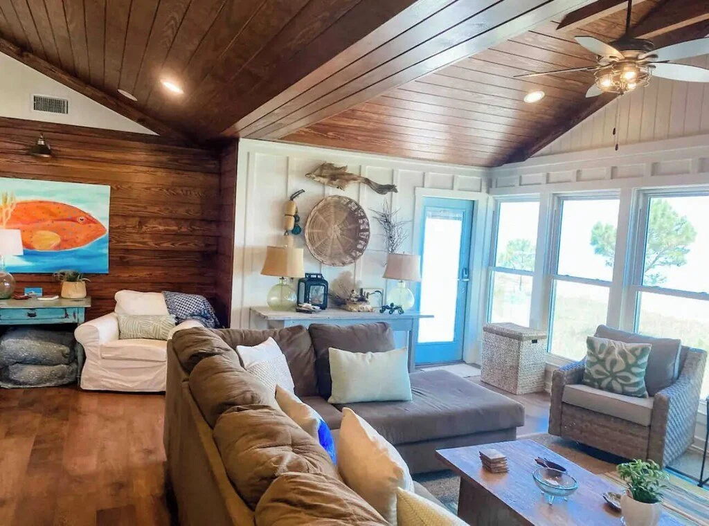 Cozy living room with wooden ceiling, large windows, beige and white furniture, and coastal decor accents.