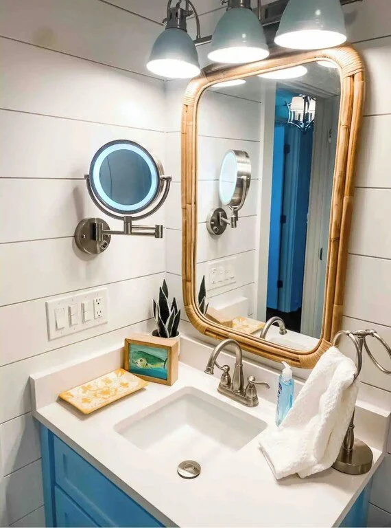 Bathroom with white shiplap walls, a large oval mirror with a bamboo frame, three overhead lights, a small round magnifying mirror, a blue vanity with a white countertop, a framed fish picture, a potted plant, a folded white towel, a soap dispenser,