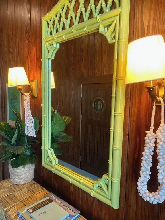A mirror with a frame made of green bamboo poles, hanging on a wooden wall in a cozy room. There are two wall-mounted lamps with yellow shades on either side of the mirror, and a large potted plant with broad leaves next to a wicker bench with magazi