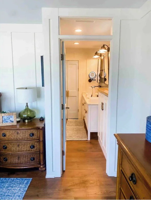 View into a small bathroom through a doorway, with white walls, a sink, mirror, and wall-mounted lights. Furniture on either side in the foreground, including a rattan dresser with a lamp and a wooden dresser.