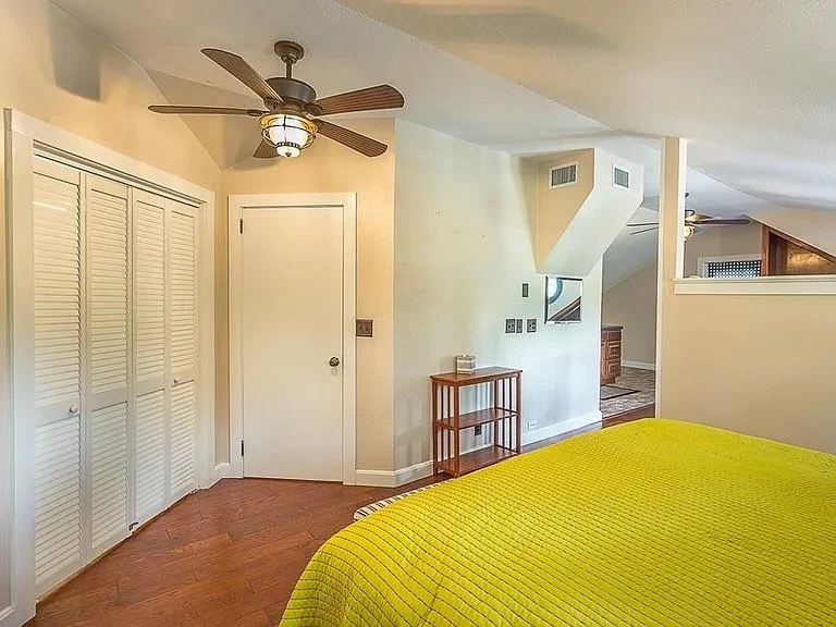Bedroom with a yellow bedspread, white louvered closet doors, ceiling fan, and small wooden shelf with decorative items.
