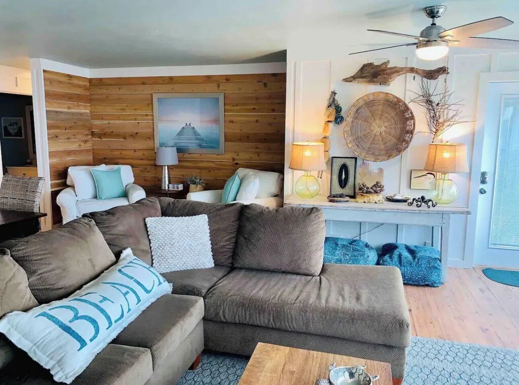 Living room with a beige sofa, white armchairs, and coastal decor including lamps, wall art, and natural wood accents.