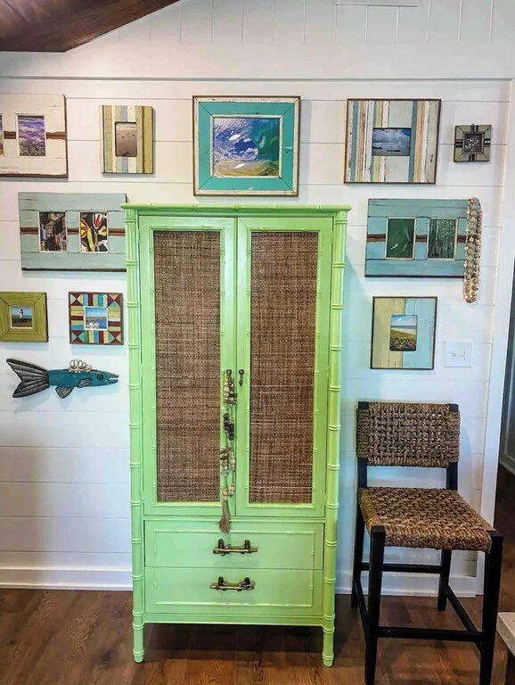 Interior of a room with a light green cabinet featuring woven doors and two drawers, surrounded by framed artwork on a white wall, and a woven chair with dark wood legs to the right.