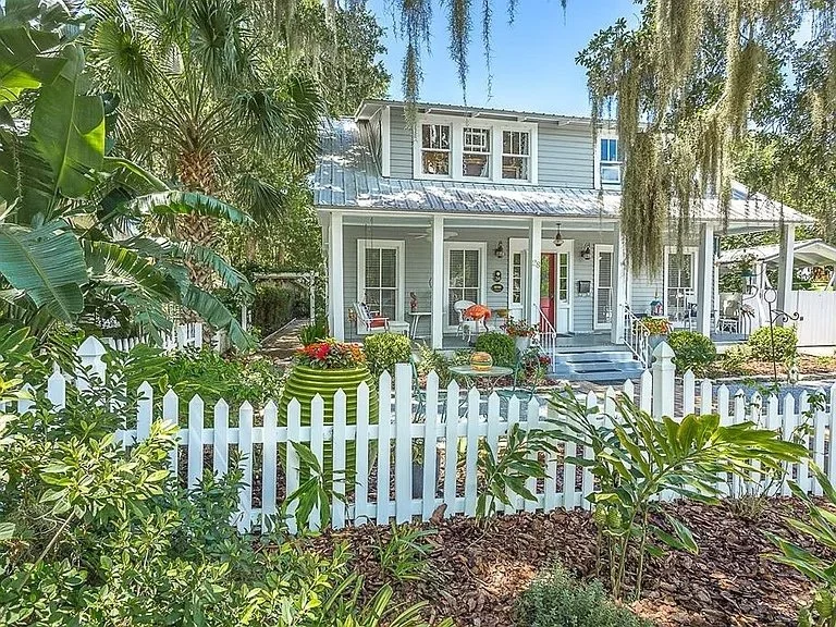 White two-story house with a front porch, surrounded by a white picket fence and lush greenery.