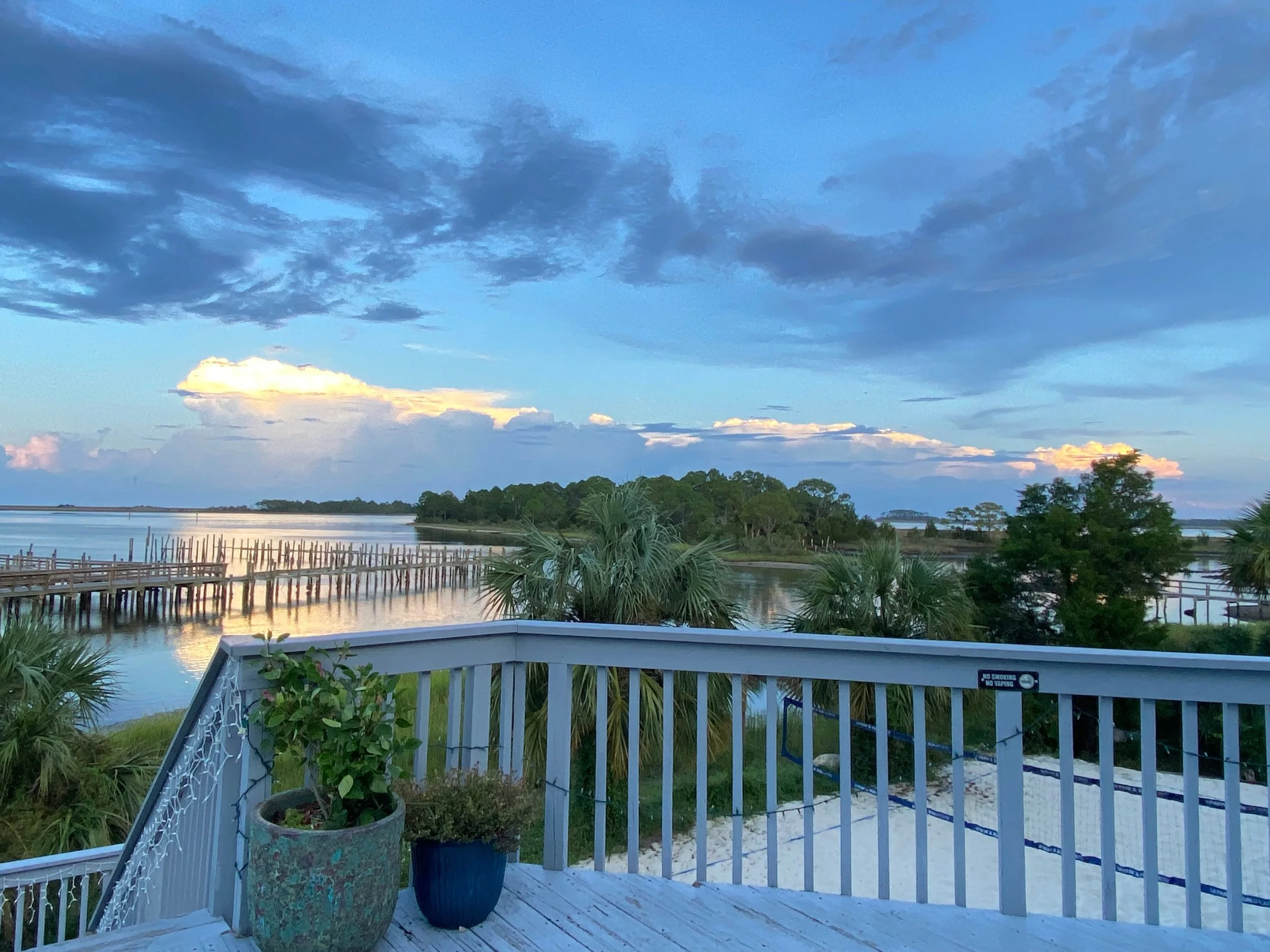View from a deck overlooking a body of water with dock, trees, and cloudy sky during sunset or sunrise.