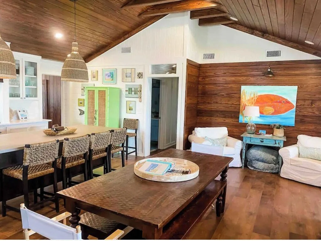 Living room with wood-paneled vaulted ceiling, wooden floor, white and green walls, wicker pendant lights, and a mix of white and green furniture including armchairs, a large wood dining table with chairs, and a blue side table with a lamp and painti