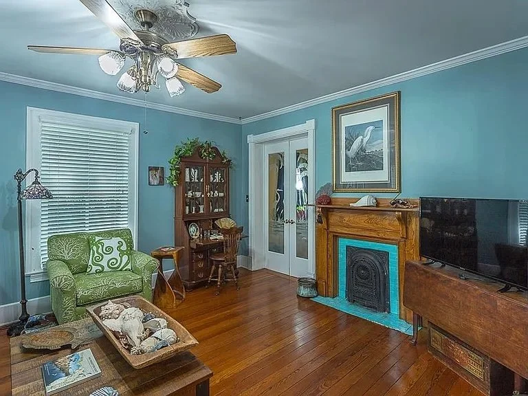 Living room with blue walls, hardwood floor, a ceiling fan, green armchair, wooden coffee table, decorative fireplace, flat-screen TV, and a wooden cabinet with glass doors.