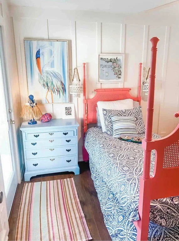 A small bedroom with a coral-colored four-poster bed, patterned bedding, and a blue dresser. Artwork on the wall includes a large painting of a stork and a framed map of Florida. A striped rug covers part of the wooden floor.